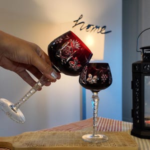 Puede incluir: Dos copas de vino de cristal rojo rubí con intrincados diseños florales y geométricos. Las copas tienen tallos largos y transparentes y están sobre una tabla de cortar de madera. La palabra "home" es visible en el fondo.