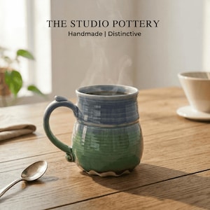 Peut inclure: Une tasse en céramique avec une partie supérieure bleue et une partie inférieure verte, dotée d'une anse incurvée et d'une base ondulée. De la vapeur s'échappe de la tasse, marquée du texte THE STUDIO POTTERY Handmade | Distinctive.