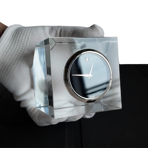 May include: A clear, square crystal clock with a black face and silver hands. The clock face has a single diamond marker at the 12 o'clock position. The clock is held in a white glove.