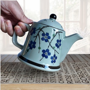May include: A pale blue ceramic teapot with a black handle and lid. The teapot features a design of blue flowers and black branches. The teapot is held above a wooden surface with wine names.