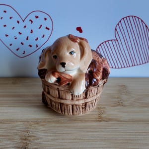 Puede incluir: Figura de cachorro de cerámica en un barril de madera. El cachorro tiene pelaje marrón y marrón, con ojos azules. El barril es de color marrón claro con líneas verticales. El fondo presenta dos contornos de corazón rojos.