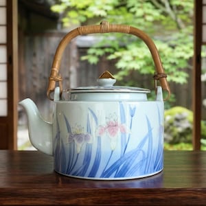 Könnte beinhalten: Eine Keramik-Teekanne mit Bambusgriff und Deckel. Die Teekanne ist mit blauen, lila und rosa floralen Designs verziert. Sie hat einen weißen Ausguss und einen goldfarbenen Akzent auf dem Deckel. Sie steht auf einer Holzoberfläche.