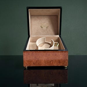 May include: Open wooden watch box with a burl wood exterior and black trim. The interior is beige with a watch holder and a gold-coloured logo. The box sits on small gold feet.