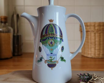 Może przedstawiać: Biały ceramiczny dzbanek do kawy z motywem balonu na ogrzane powietrze w kolorze turkusowym, zielonym i granatowym. Dzbanek ma zakrzywiony uchwyt, dzióbek i pokrywkę z małym detalem korony. Na drewnianej powierzchni.