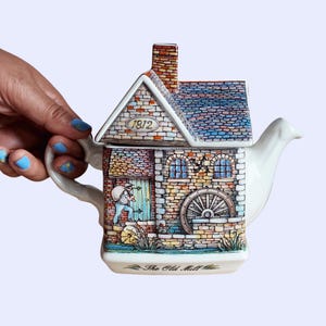 May include: A ceramic teapot shaped like a house with a water wheel, featuring a colourful illustration of a person carrying a sack near a door. The roof is tiled, the chimney is brick, and the text 1812 and The Old Mill are printed on the surface.