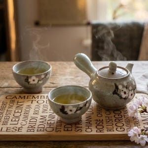 Peut inclure: Un service à thé en céramique avec une théière et deux tasses, chacune ornée d'un motif floral. La théière a une anse incurvée et un couvercle. Les tasses et la théière sont sur un plateau en bois avec du texte. De la vapeur s'élève du thé.