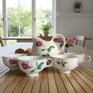 Op de afbeelding: Een crèmekleurige theeset met een kan en vier kopjes, elk versierd met een roze roos en groene bladeren. De set is op een houten tafel geplaatst, wat een klassieke en elegante uitstraling creëert. De kan is ongeveer 20 cm hoog.