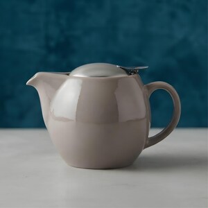 Può includere: Una teiera in ceramica color tortora chiaro con corpo arrotondato e bombato, beccuccio curvo e manico abbinato. La teiera presenta una finitura liscia e lucida ed è sormontata da un coperchio in metallo argentato lucido con una piccola linguetta.