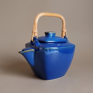 May include: A deep blue ceramic teapot with a square-shaped body, a matching lid, and a curved spout. The teapot features a light-coloured, arched bamboo handle attached to two ceramic loops on the top, providing a contrast to the glossy blue finish.