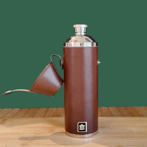 May include: A cylindrical brown leather-wrapped flask with a polished silver-tone metal cap and neck. The flask features a matching brown leather cap cover attached by a hinge. A small white square label with black text is centred near the base.