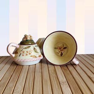 Könnte beinhalten: Dekoratives Tee-Set mit Teekanne und Tasse. Die Teekanne ist cremefarben mit floralen Designs, einem grünen Deckel und einem rosa Sockel. Die Tasse ist cremefarben mit einem floralen Design im Inneren und einem weinroten Rand.