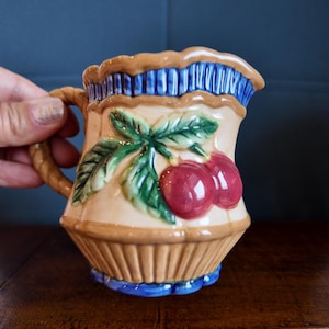 May include: A ceramic jug with a tan base, brown rim, and blue accents. The jug features a handle and is decorated with a pair of red cherries and green leaves. The jug is approximately 13 cm tall.