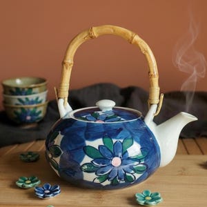 Peut inclure: Une théière en céramique avec un motif floral bleu et vert, un couvercle blanc et une anse en bambou courbée. La théière présente un bec verseur blanc et est ornée de fleurs peintes à la main. De petites pièces en céramique assorties sont posées.
