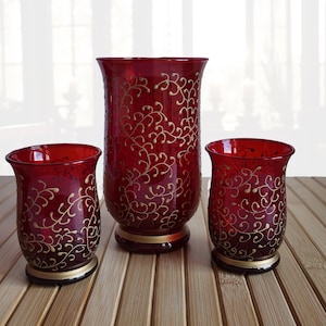 Peut inclure: Ensemble de trois vases en verre rouge ornés de motifs de vigne dorés. L'ensemble comprend un grand vase central et deux plus petits. Chaque vase est doté d'une bande dorée à la base.