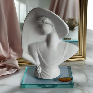 Könnte beinhalten: Eine weiße Büstenskulptur einer Frau mit breitkrempigem Hut. Die Skulptur ist auf einem transparenten, rechteckigen Glassockel montiert. Die Büste ist ein klassisches Kunstwerk mit einem ruhigen Ausdruck und eleganten Linien.