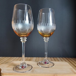 May include: Two clear glass wine glasses with amber-tinted bases and etched vertical white line patterns around the bowls. Each glass features a slender stem and a small, spherical red and white striped bead accent attached to the circular base.