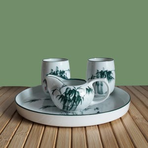 Peut inclure: Service à thé en céramique blanche avec un motif de bambou vert. L'ensemble comprend un plateau, une crémière et deux tasses. Le plateau et les tasses ont un bord vert. Le motif de bambou est peint en nuances de vert.