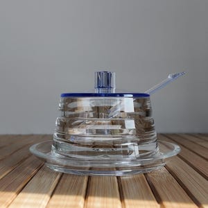 Peut inclure: Un sucrier en verre transparent avec un bord et un couvercle bleus, et une cuillère assortie. Le bol repose sur une soucoupe en verre transparent. Le design présente des rainures horizontales. Le fond est gris clair.