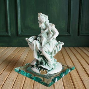 Könnte beinhalten: Eine weiß-grüne Keramikfigur einer Mutter und eines Kindes mit goldenen Akzenten, die auf einer klaren, quadratischen, grün umrandeten Glasschale sitzt. Die Figur ist mit fließenden Gewändern und einem ruhigen Ausdruck versehen. Die Glasschale ist etwa 13 cm groß.