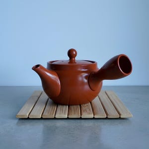 Könnte beinhalten: Eine braune Keramik-Teekanne mit seitlichem Griff und kleinem Ausguss. Die Teekanne steht auf einem Bambus-Topfuntersetzer. Die Teekanne hat einen abgerundeten Körper und einen Deckel mit einem kleinen Knopf. Der Hintergrund ist hellblau.