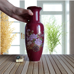 May include: A burgundy ceramic vase with a floral design. The vase features a wide base, a slender body, and a flared rim with a gold trim. The floral design includes purple and white peonies and other flowers. The vase is approximately 12 inches tall.