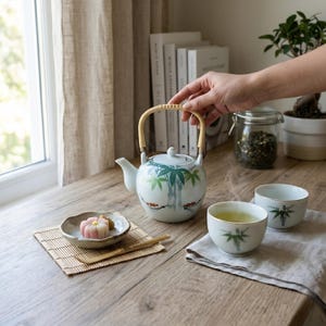 May include: A white ceramic teapot with a bamboo handle and a bamboo and green tree design. Two matching teacups with bamboo designs are on a wooden table. A small plate with a pink and white dessert is on a bamboo mat.