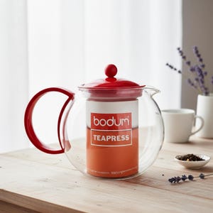 Puede incluir: Una tetera Bodum de vidrio transparente con tapa, asa y émbolo rojos. La tetera tiene un interior cilíndrico y está llena de líquido naranja. El logotipo de Bodum y "TEAPRESS" están impresos en el lateral.