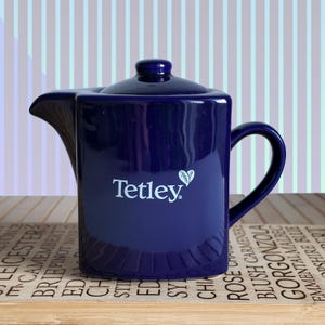 Puede incluir: Una tetera de cerámica azul marino con tapa y asa. El logotipo de Tetley está en blanco en la parte delantera. La tetera está sobre una bandeja de madera con texto.