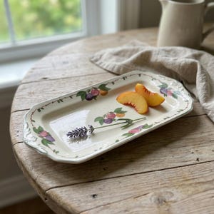 May include: Rectangular off-white serving tray with a floral and fruit design. The tray has a green border and is decorated with illustrations of plums, peaches, and leaves. Two peach slices and a sprig of lavender are on the tray.