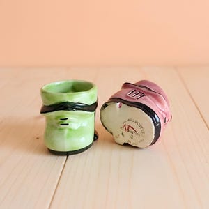 以下が含まれることがあります： 靴の形をした陶器のオーナメント2点。1点は黒い縁取りと横線が入ったライトグリーン。もう1点は黒い縁取りと窓のグラフィックが施されたピンク色で、底面にはRoyal Art Pottery, Longton, C, Made in Englandと刻印されています。