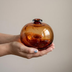 May include: A spherical, amber-toned glass vase with a small opening and a dark rim. The vase has a bubble-like texture and is held in cupped hands. The glass reflects light, creating highlights and shadows. The vase is a decorative item.