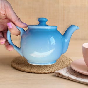 May include: A glossy, light blue ceramic teapot with a rounded body, a curved handle, a matching lid with a small knob, and a tapered spout. The teapot features a smooth, reflective finish that transitions from a deeper blue to a lighter shade.