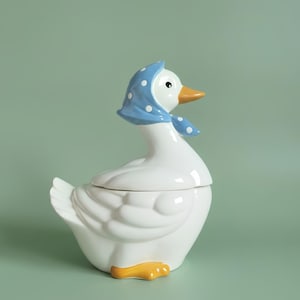 May include: A white ceramic goose figurine featuring a glossy finish, a bright orange beak, and orange feet. The goose wears a light blue headscarf decorated with small white polka dots tied under its chin, with a black eye visible on the side.