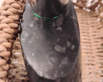 Puede incluir: Primer plano de un objeto gris oscuro con superficie texturizada. Una fina línea verde brillante es visible cerca de la parte superior. El objeto está parcialmente oscurecido por un elemento metálico brillante. El fondo presenta un material tejido de color marrón claro.