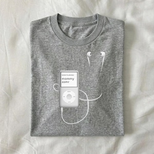 May include: A heather gray t-shirt featuring a white iPod with the text "NOW PLAYING: mommy asmr" on the screen. White earbuds are positioned near the collar of the shirt, with the cord connected to the iPod.