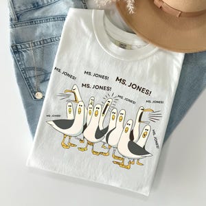 May include: White t-shirt with a cartoon of seagulls and the text "MS. JONES!". The seagulls have black and grey wings and yellow feet. The shirt is shown with blue jeans and a tan hat. The shirt is made of a soft material.