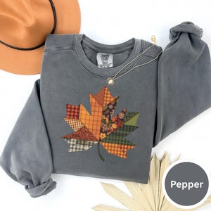Puede incluir: Una sudadera gris con un diseño de hoja de patchwork en colores otoñales. La hoja está compuesta por varios patrones de tela en tonos naranja, rojo y verde. También se ve un collar dorado y un sombrero marrón.