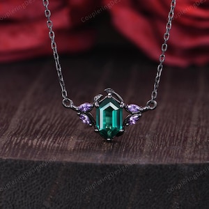 May include: A black necklace with a central emerald-cut green gemstone, flanked by smaller purple gemstones. The necklace has a delicate chain and is set against a dark wooden surface and a blurred red floral background.