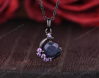 May include: A black pendant necklace featuring a dark blue gemstone with sparkling inclusions, accented by small purple gemstones. The pendant is suspended from a delicate black chain. The background is a blurred dark red.