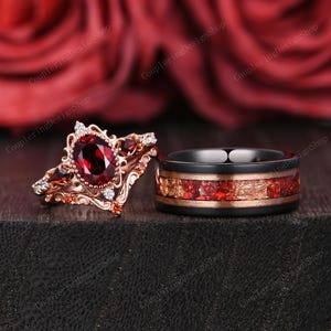 May include: A rose gold engagement ring set with a large oval-cut red gemstone and smaller accent stones. The matching wedding band has a black band with a red and gold inlay. The rings are displayed against a blurred background of red roses.