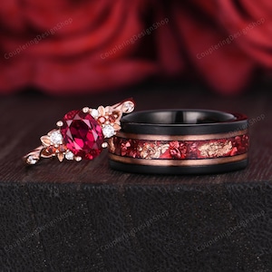 Peut inclure: Une bague de fiançailles en or rose avec un grand rubis ovale au centre, entouré de petites pierres précieuses et de motifs floraux. Une alliance noire avec des incrustations rouges et dorées est à côté.