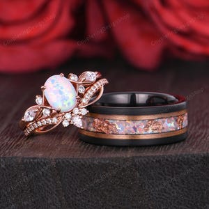 Op de afbeelding: Een roségouden verlovingsring met een ovale opaal middensteen en een bijpassende trouwring. De trouwring heeft een zwarte band met een koperkleurige inleg en opaalaccenten. De verlovingsring heeft een bloemenontwerp met kleine diamanten.