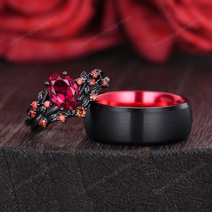 Könnte beinhalten: Ein schwarzes Hochzeitsring-Set. Ein Ring hat einen großen ovalen Rubin-Edelstein und florale Akzente. Der andere Ring ist ein massives schwarzes Band mit roter Innenseite. Die Ringe sind vor einem dunklen Hintergrund platziert.