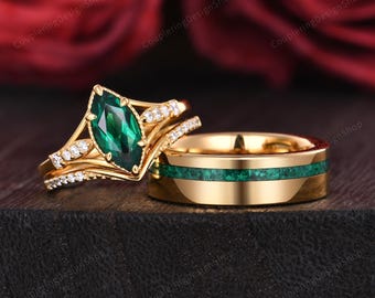May include: A gold wedding ring set featuring a marquise-cut emerald engagement ring with diamond accents and a matching band with a green inlay. The rings are displayed on a dark surface with a blurred red background.