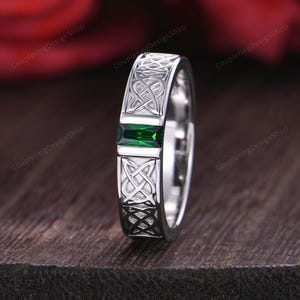 Peut inclure: Une bague en argent ornée d'un motif de nœud celtique et d'une pierre précieuse rectangulaire vert émeraude. La bague est présentée sur une surface en bois sombre. Le texte "CouplerRingDesignShop" est visible.