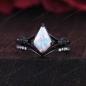 May include: A black engagement ring set featuring a kite-shaped opal center stone. The ring is accented with small green gemstones and a band with white gemstones. The set is designed with a unique, geometric style.