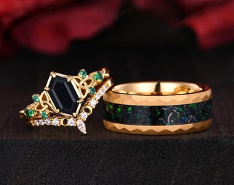 May include: A set of two gold wedding bands. The engagement ring features a black octagon-shaped gemstone surrounded by smaller green gemstones and a row of smaller white gemstones. The wedding band is a gold band with a black and green inlay.