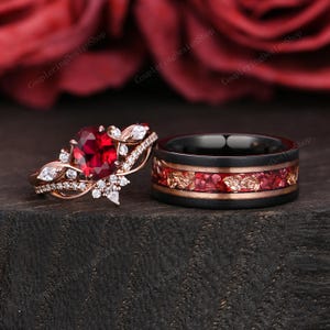 May include: A rose gold engagement ring with a heart-shaped red gemstone and a matching black wedding band. The wedding band has a red and gold inlay design. Both rings are set against a dark background.