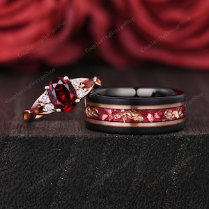 May include: A rose gold engagement ring featuring a central oval-cut deep red garnet flanked by smaller clear stones and marquise-cut red stones, paired with a black band featuring a central inlay of crushed red stone and gold-colored flakes.