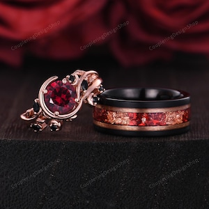 Peut inclure: Deux bagues, l'une avec une grosse pierre rouge ronde et des accents noirs, l'autre une bande noire avec une incrustation rouge et or. Les bagues sont posées sur un fond sombre, avec le texte "CoupleringDesignShop" visible.